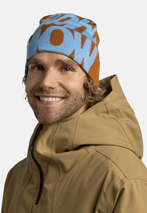 Knit beanie with a light blue base and brown abstract patterns; soft texture; worn with a tan hooded jacket featuring a zipper detail.