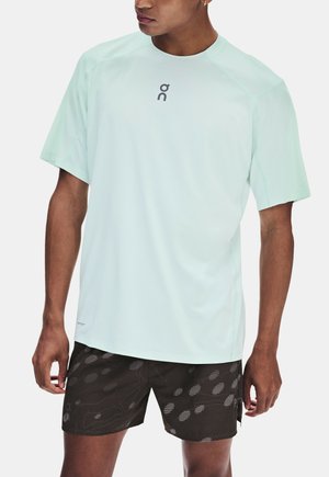 Uomo con maglietta sportiva azzurro chiaro a maniche corte e pantaloncini neri con motivo, in piedi con le braccia rilassate lungo i fianchi.