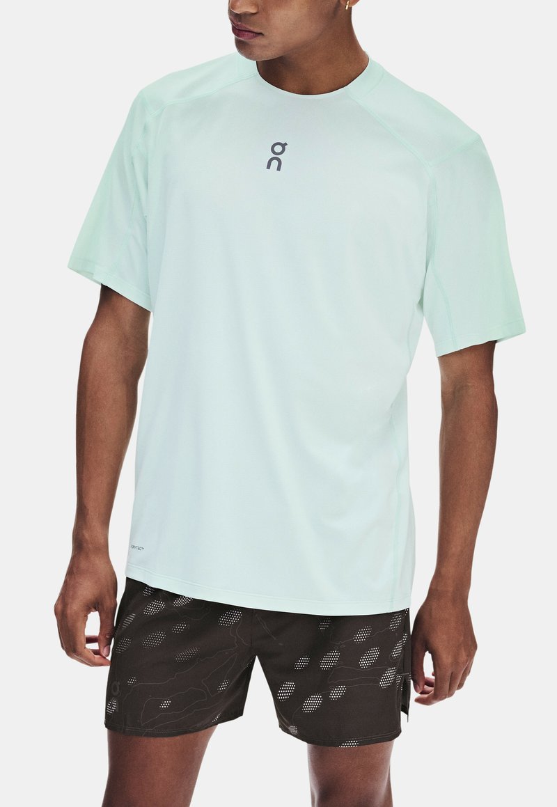 Homme portant un t-shirt de sport bleu clair à manches courtes et un short noir à motifs, debout avec les bras détendus le long du corps.