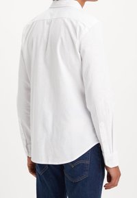 Camicia bianca a maniche lunghe in cotone, con colletto a puntale, orlo curvo e polsini con un bottone, indossata con jeans blu.