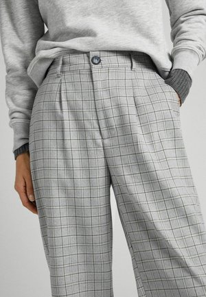 Person iført grå ternede bukser med høj talje, plisseringer og en knap, kombineret med lagdelte grå trøjer, hånd i lommen.