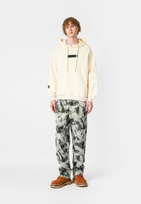 Sudadera crema con cordón ajustable, acento negro en el pecho. Pantalones verdes y blancos con patrón, combinados con zapatos de ante marrón.