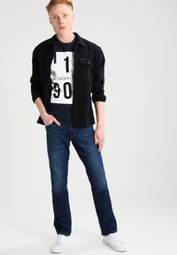 Camisa negra de botones sobre una camiseta gráfica azul marino con texto blanco, combinada con jeans azul oscuro y zapatillas blancas.