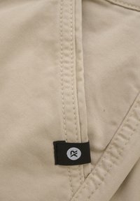 Beige stof med synlige sømme og et sort mærke med en hvid cirkel og et "R" på kanten. Teksturen ser glat og let ud.