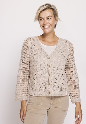 Lächelnde Frau, die eine beige gehäkelte Strickjacke mit Blumenmuster und beige Hose trägt, steht vor einem schlichten hellen Hintergrund.
