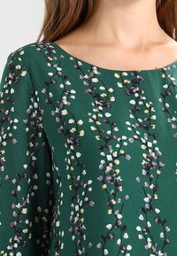 Vestido floral verde con un escote redondo, con un delicado patrón de flores blancas, amarillas y moradas sobre una textura de tela suave.