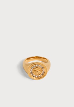 Guldring med en rund front, der har et hævet løvehoved-design omgivet af en cirkel af klare ædelstene på hvid baggrund.