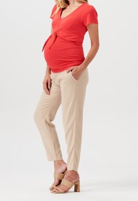 Maglietta premaman rossa aderente con scollatura a V e dettaglio con fiocco, abbinata a pantaloni beige a sigaretta. Il modello indossa sandali con tacco quadrato nudi.