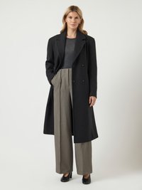 Naine, kes kannab pikka musta mantlit tumeda särke ja laiade beežide pükstega, seisab ühe käega taskus, tasase tausta ees.