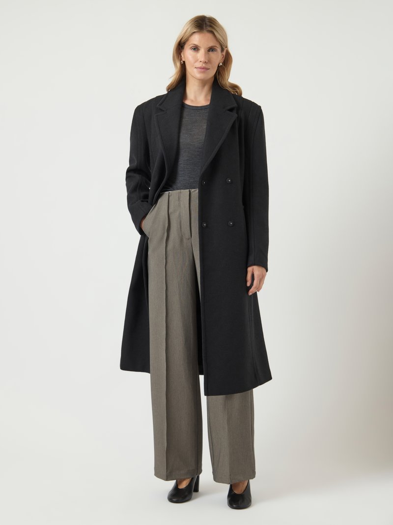 Naine, kes kannab pikka musta mantlit tumeda särke ja laiade beežide pükstega, seisab ühe käega taskus, tasase tausta ees.