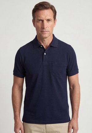 Man draagt een donkerblauw poloshirt met korte mouwen en een borstzakje en een beige broek, staand tegen een effen lichte achtergrond.