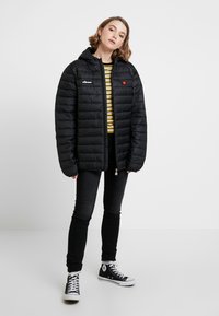 Doudoune noire avec capuche, présentant un zip sur le devant et un écusson de marque sur la poitrine. Portée par-dessus une chemise rayée avec un jean noir et des baskets.