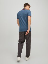 Mann iført blå polo skjorte og mørke bukser, står med ryggen til kameraet på en ensfarget lys bakgrunn.