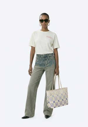 Žena v bílem tričku Diesel, široké vybledlé džíny, černé špičaté boty, sluneční brýle, kruhové náušnice, drží béžovou tašku s modrými vzory.