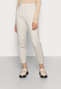 Beige maßgeschneiderte Hose mit schmaler Passform, ausgestattet mit Vordernähten, Seitentaschen und einer glatten Textur. Kombiniert mit beigefarbenen Loafers und schwarzer Sohle.