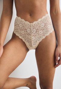 Intimo in pizzo di colore nude con una cintura ondulata, motivo floreale e un pannello posteriore liscio. Presenta bordi e dettagli delicati.