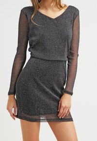 Mustat kimalteet mekko, jossa on pitkät verkkohihat, V-pääntie, vartalonmyötäinen vyötärö ja mini pituus. Kimalteleva pinta lisää visuaalista kiinnostavuutta.