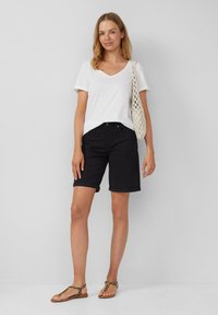 Shorts en denim noir avec des revers, assortis à un t-shirt blanc à col en V. Le modèle porte un sac en toile crocheté et des sandales plates à lanières.