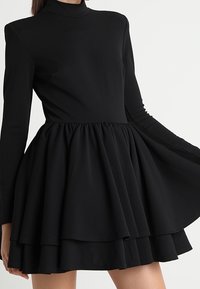 Mustat mekko korkealla kauluksella, pitkähihaisilla anteilla ja liehuvalla, kerroksittain leikatulla hameella. Valmistettu sileästä kankaasta, jossa on rypistetty vyötärö.