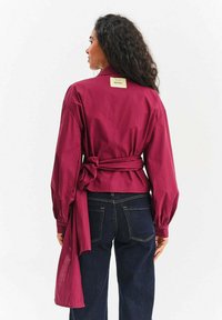 Bordowa kopertowa koszula z długimi rękawami, wiązana w talii; wykonana z miękkiego materiału z metką na karku z tyłu. W połączeniu z ciemnymi dżinsami.