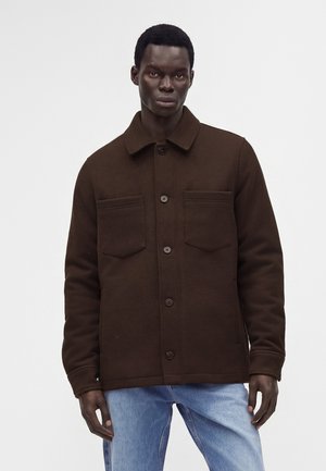 Homme portant une veste boutonnée marron foncé avec deux poches sur la poitrine et un jean bleu clair, debout devant un fond uni clair.