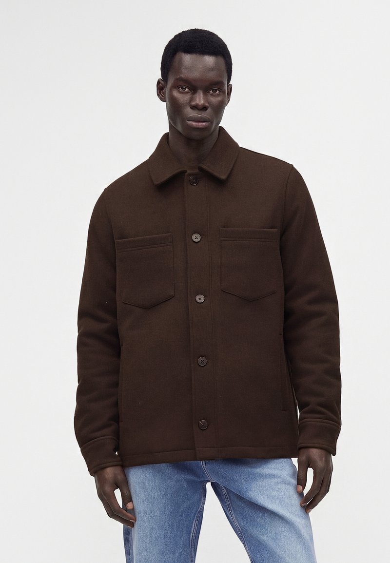 Homme portant une veste boutonnée marron foncé avec deux poches sur la poitrine et un jean bleu clair, debout devant un fond uni clair.