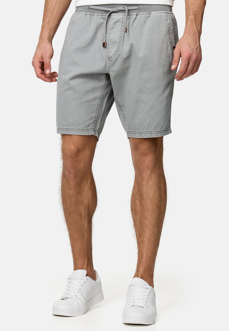 Homme portant un short gris clair à cordon et des baskets blanches, debout les jambes légèrement écartées devant un fond uni.