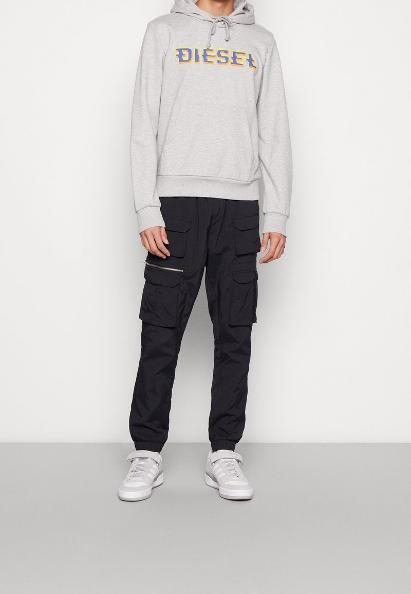 Homme portant un sweat à capuche Diesel gris clair et un pantalon cargo noir à multiples poches, associé à des baskets blanches, debout contre un mur blanc.