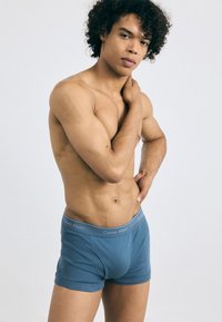 Boxeri din bumbac albastru cu un elastic gri pe care este brandul "Calvin Klein". Modelul stă cu o expresie neutră, evidențiind croiala.
