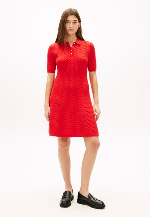 Žena stojící, oblečená v červených texturovaných pletených šatech po kolena s krátkými rukávy a černými mokasíny na bílém pozadí.