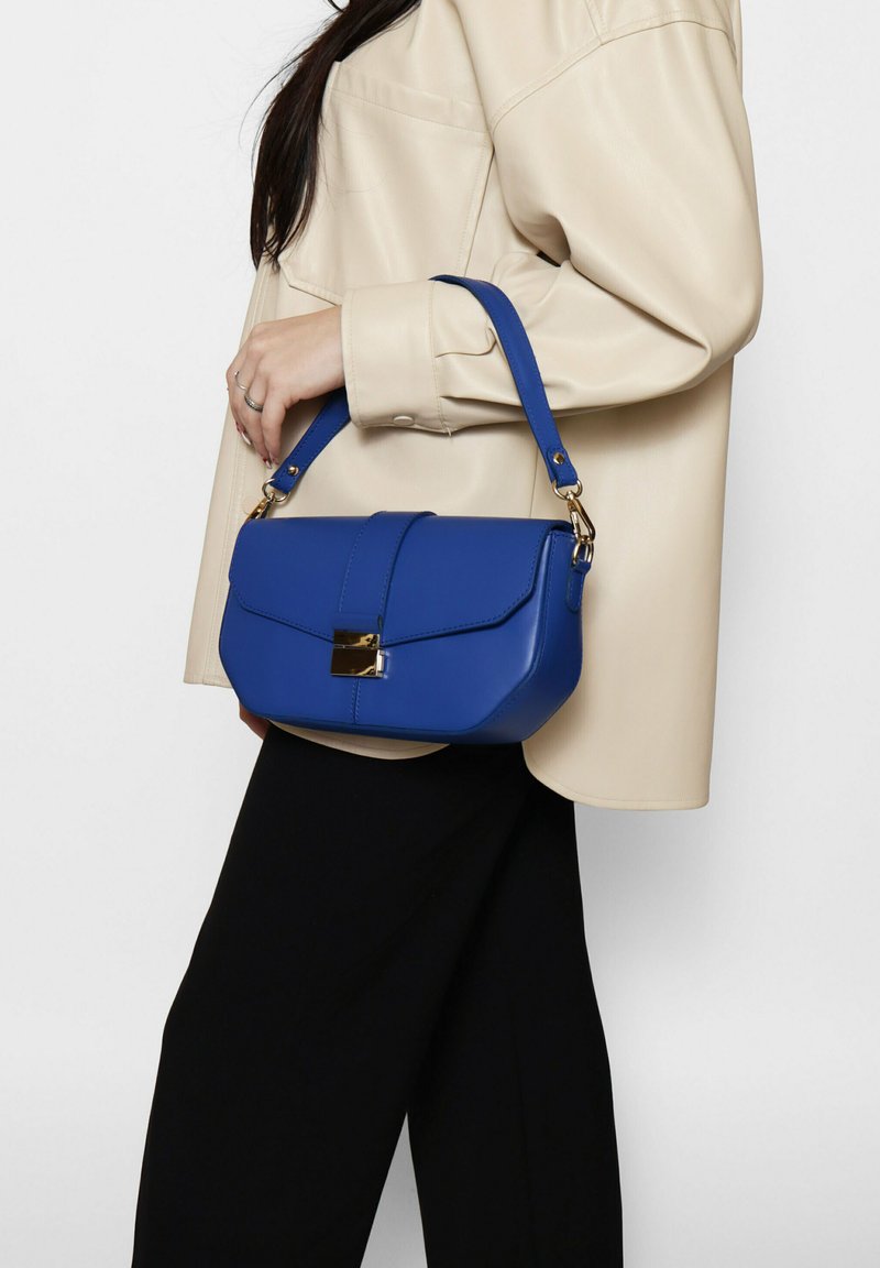 Borsa blu in pelle sintetica con una forma strutturata, chiusura a pattina, hardware dorato e manico corto. Indossata con una camicia oversize beige.