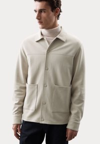 Chaqueta de un beige claro hecha de un material suave, con cierre de botones, grandes bolsillos frontales, cuello de camisa y detalles de costura en contraste.