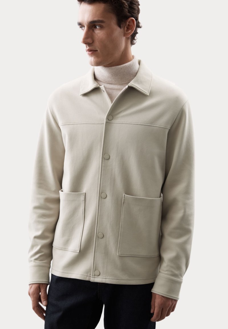 Chaqueta de un beige claro hecha de un material suave, con cierre de botones, grandes bolsillos frontales, cuello de camisa y detalles de costura en contraste.