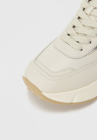 Κρεμ δερμάτινο αθλητικό παπούτσι με στρογγυλή μύτη· διαθέτει υφασμένες κορδόνια και μια λεπτή μεταλλική λεπτομέρεια. Η σόλα είναι ανοιχτό καφέ με αυλακώσεις.