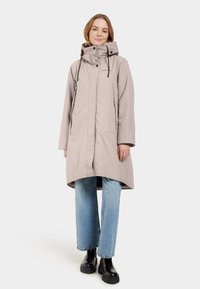 Donna che indossa un cappotto invernale a mezzaluna di colore beige chiaro con cappuccio, jeans blu chiaro e stivali neri chunky, in piedi contro uno sfondo bianco.
