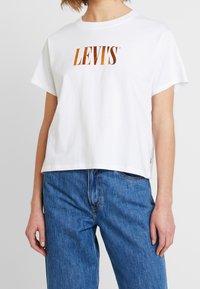 Valkoinen puuvillainen t-paita lyhyillä hihoilla, jossa on oranssi Levi's-logo rinnan kohdalla. Yhdistettynä sinisiin farkkuihin.