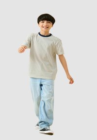 Maglietta a righe a maniche corte beige e blu navy, abbinata a jeans azzurri e sneakers nere. Il soggetto sta camminando e sorridendo.