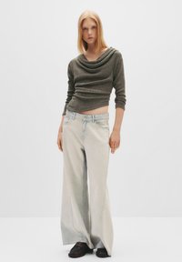 Dunkelgrüner, langärmeliger Rippenpullover mit Kapuze, kombiniert mit hellblauen weiten Jeans und schwarzen Slippern. Glatte Texturen, lässiger Stil.