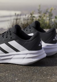 Chaussures de course Adidas noires et blanches avec semelles Cloudfoam posées à l'extérieur sur une surface texturée avec des plantes floues en arrière-plan.