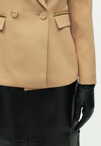 Μπεζ διπλόκλειδο σακάκι με χρυσά κουμπιά, διακοσμημένο με αλυσίδα στις τσέπες και συνδυασμένο με μαύρη δερμάτινη φούστα.