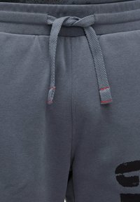 Graue Sweatpants aus einem strukturierten Stoff, mit einem Kordelzug in Rotakzenten und einem großen schwarzen Grafikdruck auf dem linken Bein.