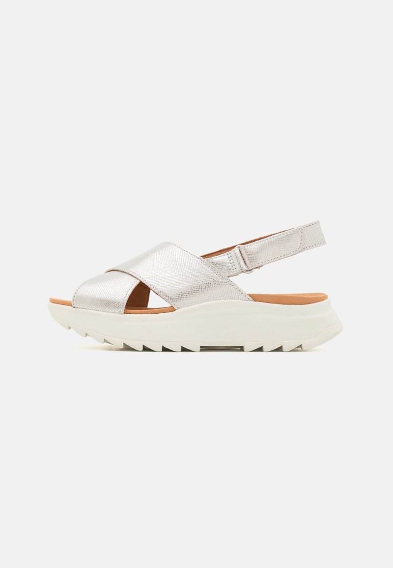 Sandale métallisée argentée pour femme avec brides croisées, bride de talon réglable, semelle beige rembourrée et semelle extérieure en caoutchouc blanche crantée.