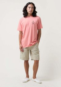 Camiseta de algodón rosa claro con un pequeño diseño gráfico negro, combinada con pantalones cortos de cargo beige y zapatillas blancas. Un atuendo sencillo y casual.