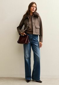 Bruine cropped jas met een bontkraag, bruine trui, wijde blauwe jeans en een kastanjebruine tas. Glanzende puntige schoenen.