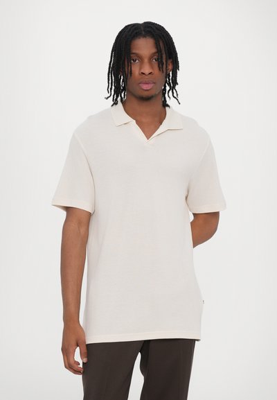 Polo shirt beige clair à manches courtes avec un col en V et un col replié, fabriqué en tissu texturé. Doté d'une coupe décontractée et de fentes sur les côtés.