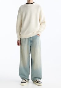 Romige gebreide trui met ribbelrand, gecombineerd met vervaagde lichtblauwe wijdere spijkerbroek en zwart-witte sneakers.