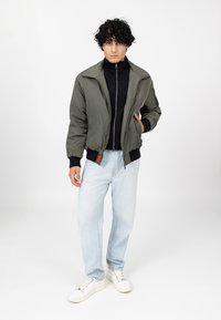 Veste bomber vert olive avec fermeture éclair sur le devant, poignets côtelés et col noir portée sur un haut noir, associée à un jean bleu clair et des baskets blanches.
