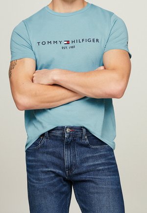 Mężczyzna noszący jasnoniebieską koszulkę Tommy Hilfiger z logo i ciemnoniebieskie dżinsy, z założonymi ramionami, beżowe tło, tatuaż na lewej ręce.
