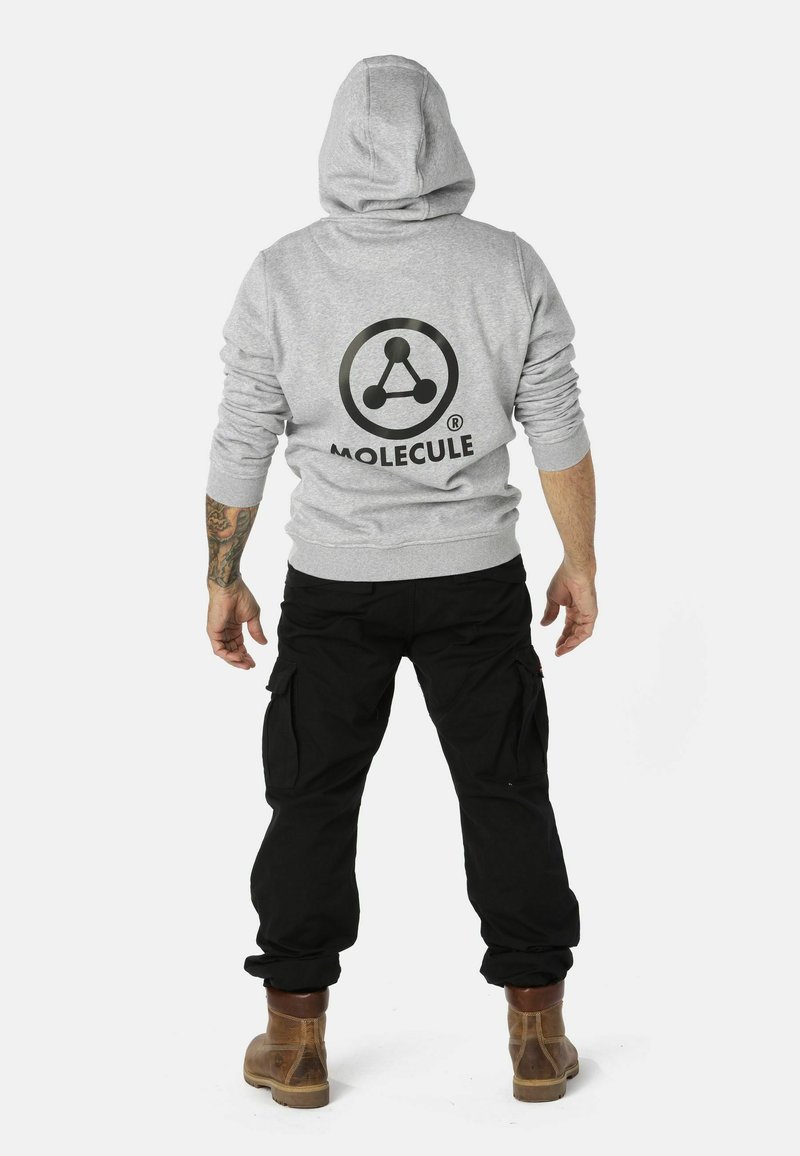 Grijze hoodie met een logo dat een cirkel en moleculaire structuur op de achterkant toont. Draag het met zwarte cargobroeken en bruine laarzen.