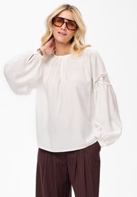 Blondynka w dużych okrągłych okularach przeciwsłonecznych w stylu żó�łwi, białej bluzce z koronkowymi detalami na rękawach i brązowych spodniach w paski, pozująca z ręką na szyi.